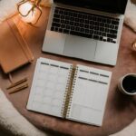 A cozy workspace features a laptop, planner, notebook, pens, two warm lights, and a coffee cup on a soft, round table.