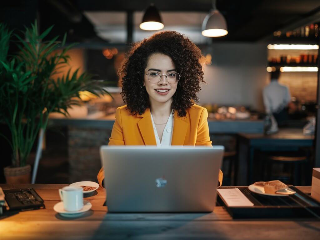 A person with curly hair and glasses uses a laptop in a cozy cafe. Coffee and pastries are present on the table.