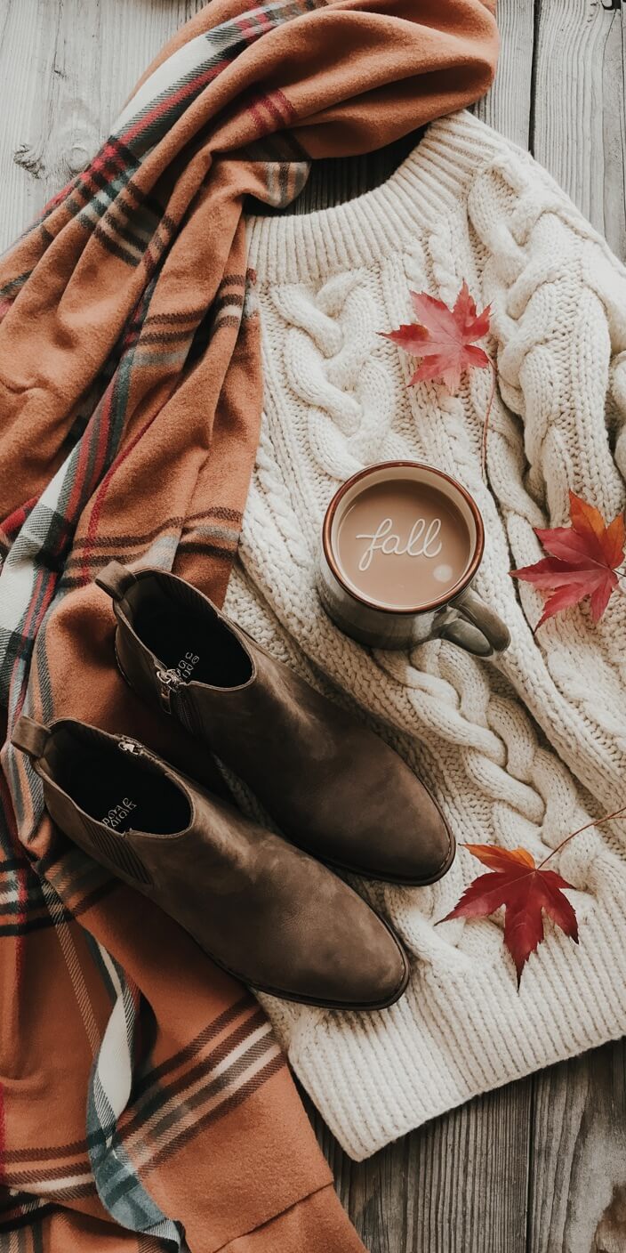 Cozy fall scene with brown boots, a cream sweater, plaid scarf, leaves, and a mug of coffee on a wooden surface.