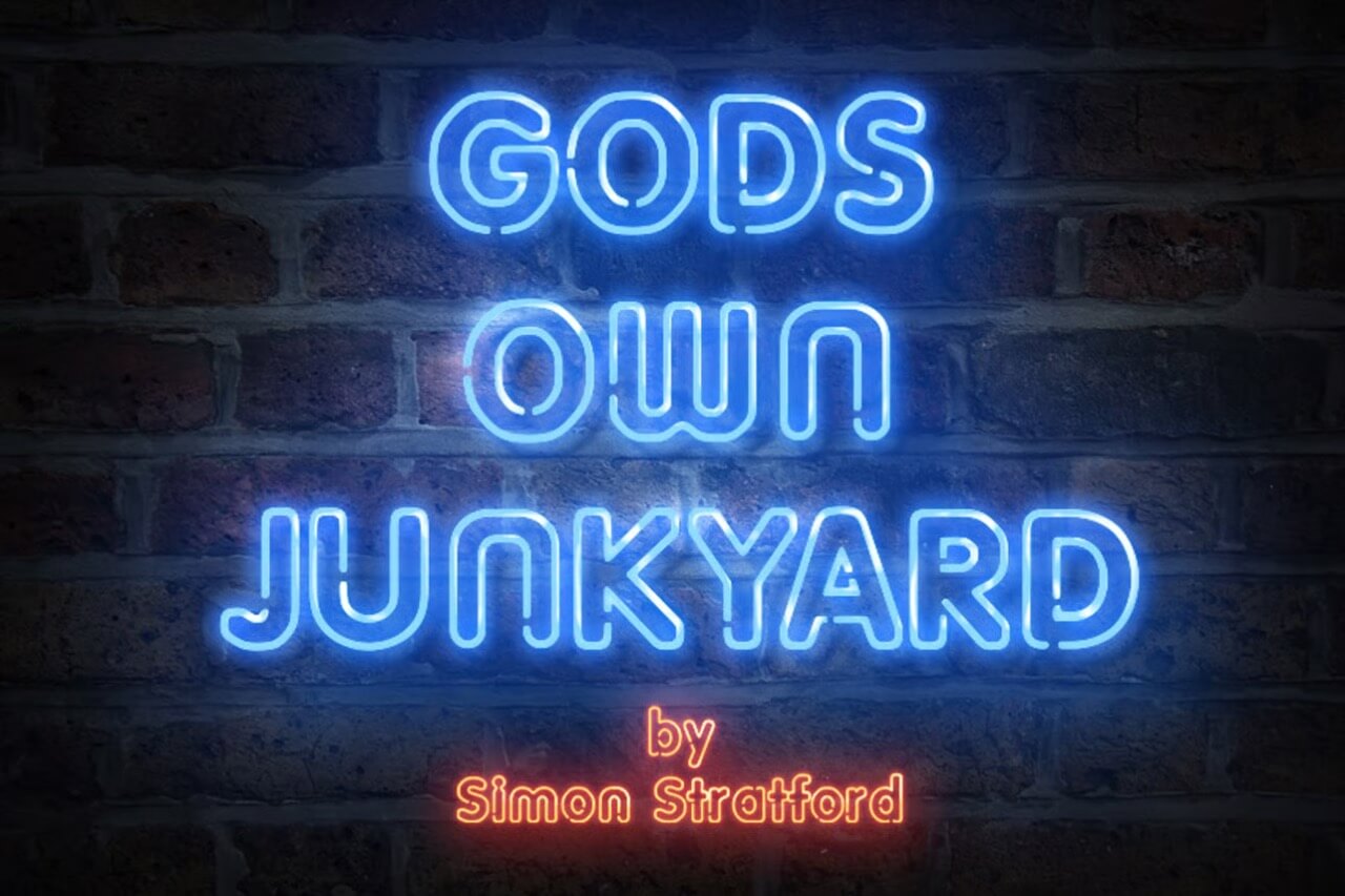 Neon sign reading "Gods Own Junkyard by Simon Stratford" on a brick wall, blue and orange glowing letters, artistic expression.
