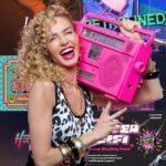 A person with curly hair and leopard-print top holds a pink boombox, surrounded by colorful, retro-themed posters and graphics.